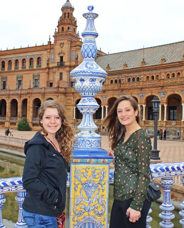 两名学生站在一座西班牙建筑前的桥上，拿着一根装饰柱子